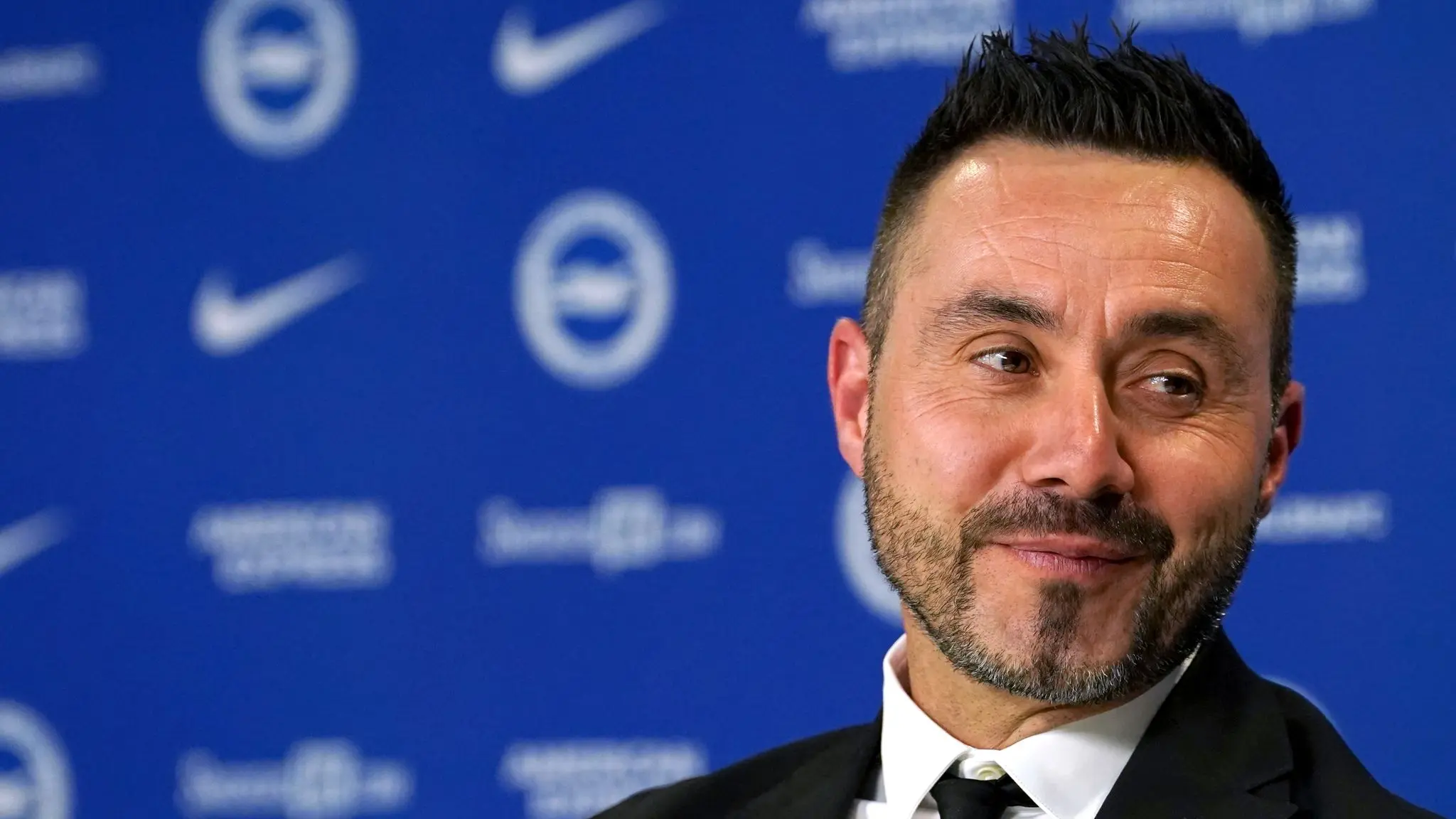 Roberto De Zerbi smiling during a press conference with Brighton & Hove Albion logos in the background