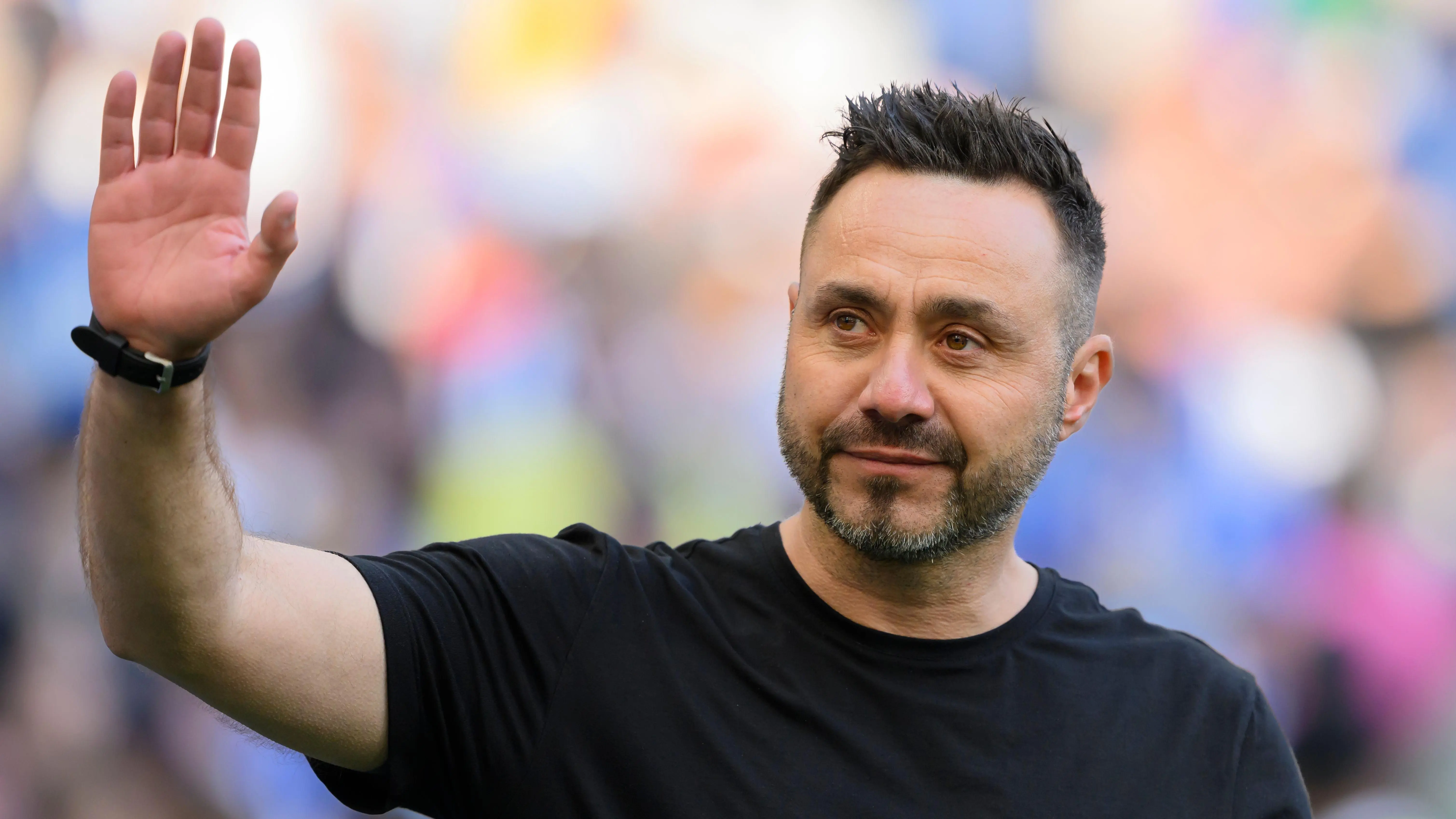 Roberto De Zerbi waving to the crowd, wearing a black shirt with a blurred background.