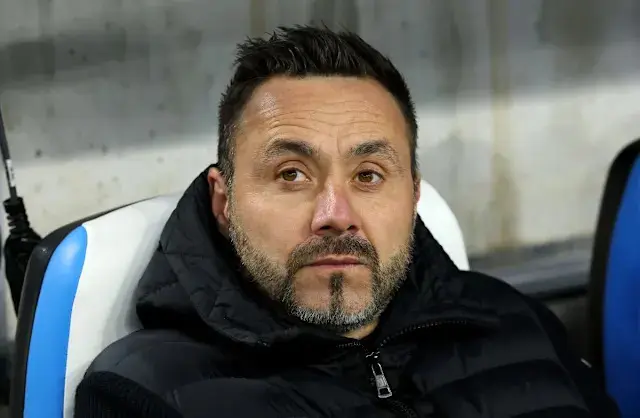 Roberto De Zerbi seated on the bench, wearing a black jacket with a serious expression.