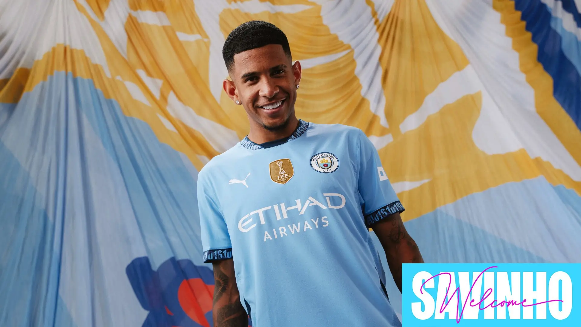 Savinho in Manchester City jersey smiling in front of colorful backdrop.