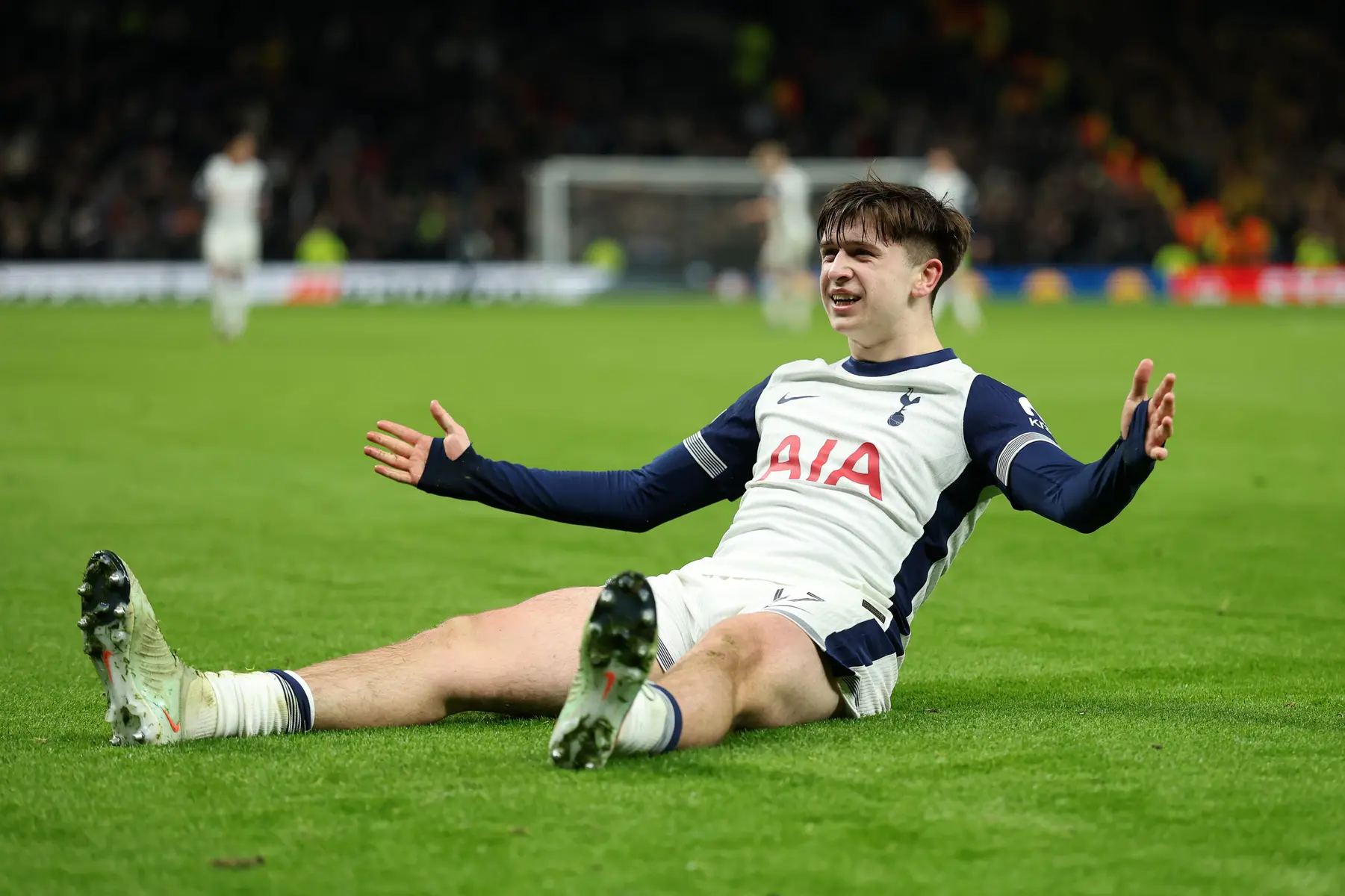 Mikey Moore in a Tottenham Hotspur kit sitting on the grass with arms outstretched during a match.