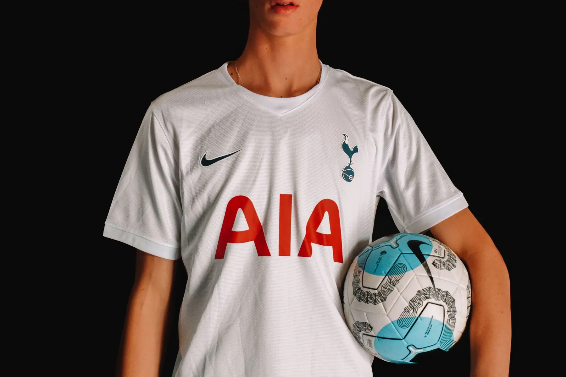 A person wearing a white Tottenham Hotspur home jersey holding a soccer ball against a black background
