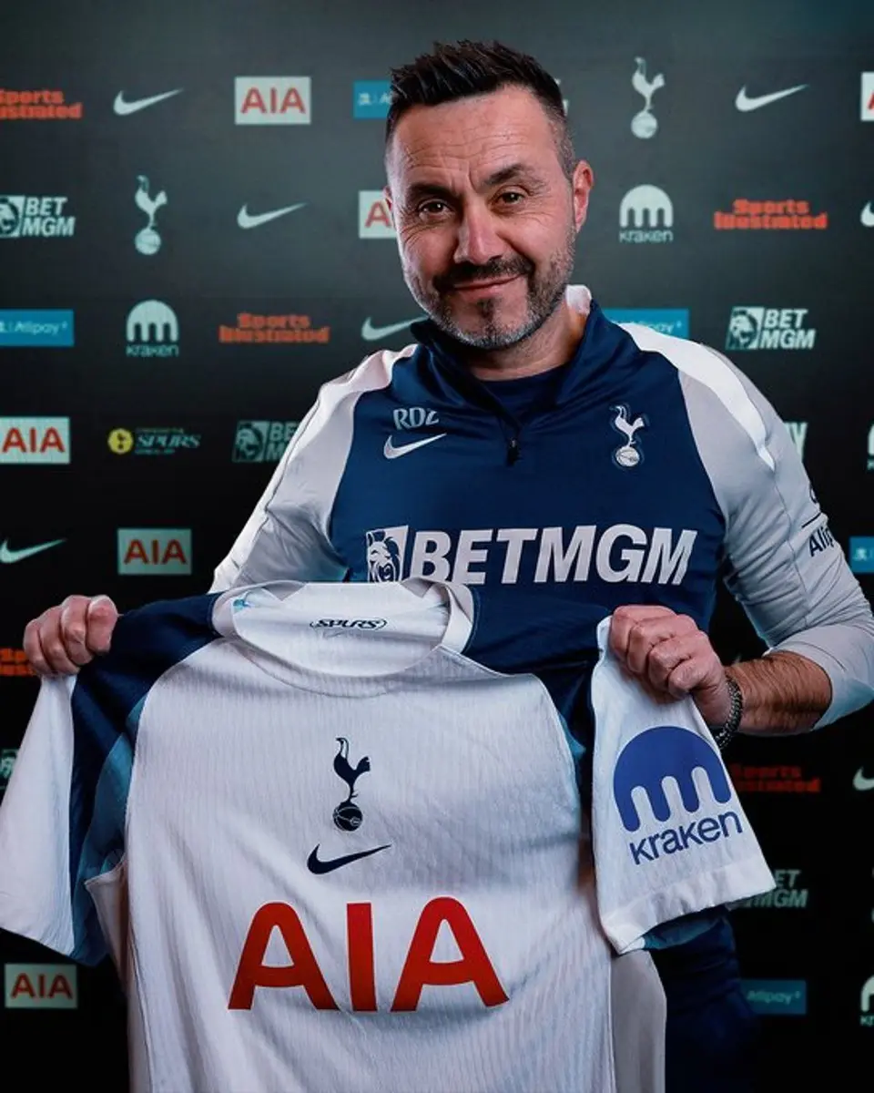 Roberto De Zerbi smiling while holding up a white Tottenham Hotspur home jersey