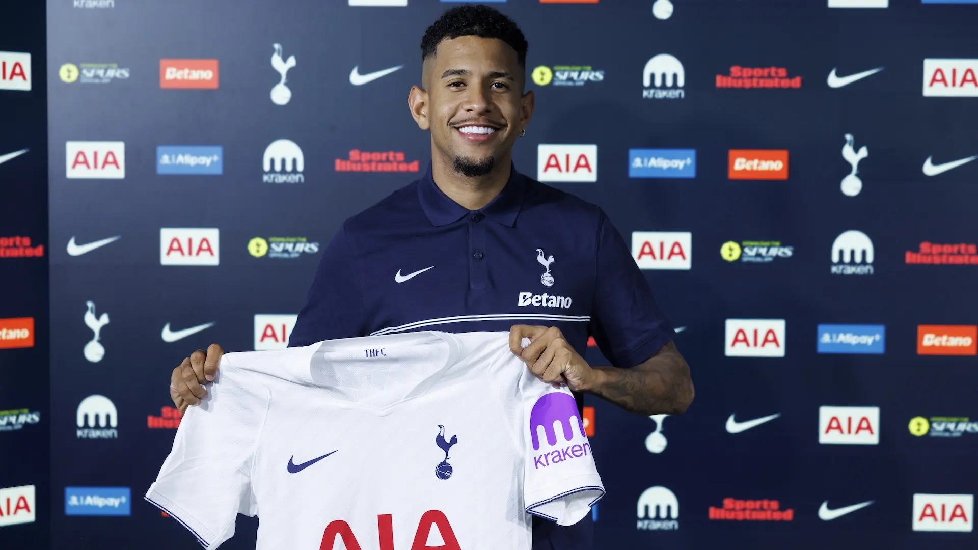 Savio smiling while holding up a white Tottenham Hotspur home jersey in front of a branded backdrop