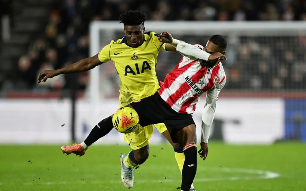 Tottenham Hotspur player in yellow AIA kit competing for the ball against a Brentford player in red and white stripes.