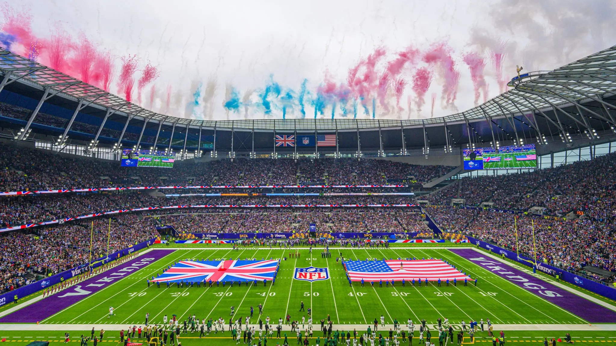 Wide view of an NFL game at Tottenham Hotspur Stadium with US and UK flags on the field and red smoke in the air