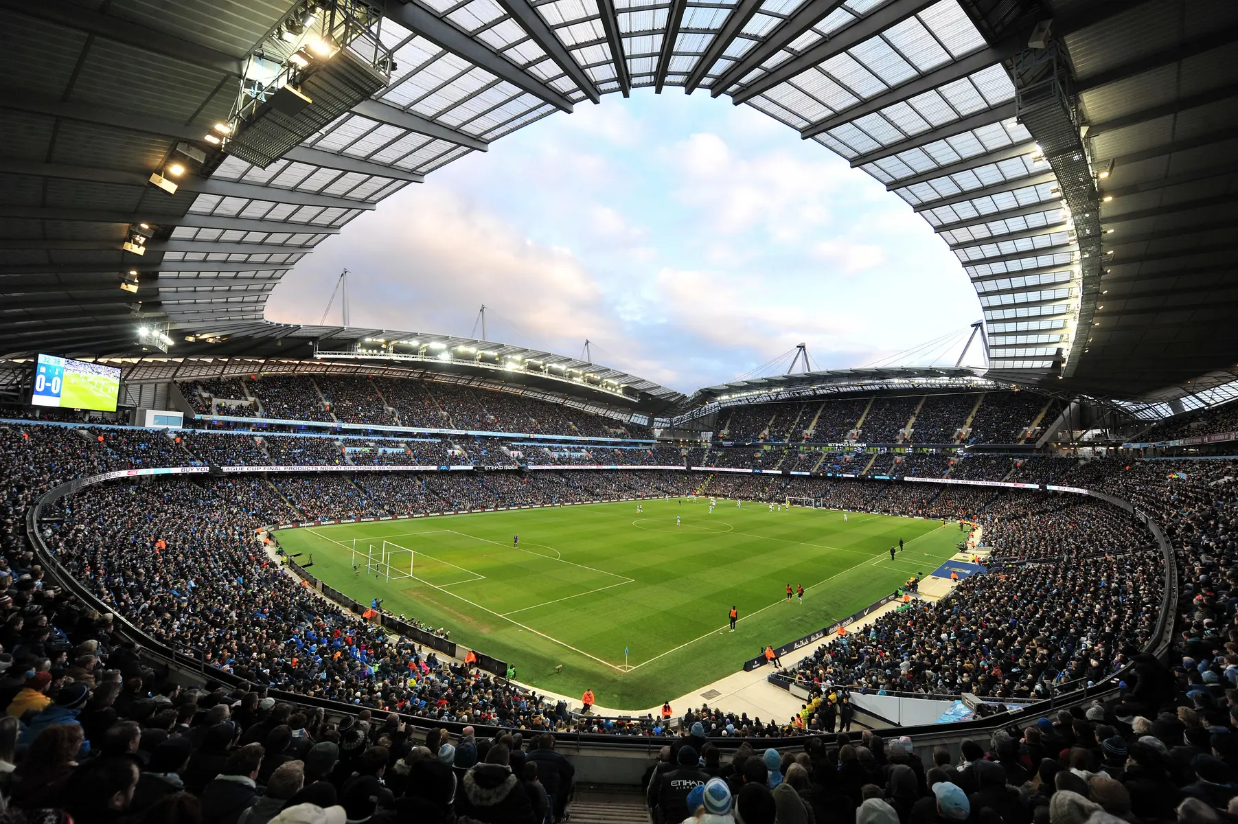 A wide-angle view of a crowded Etihad Stadium during a football match under a partially open roof.