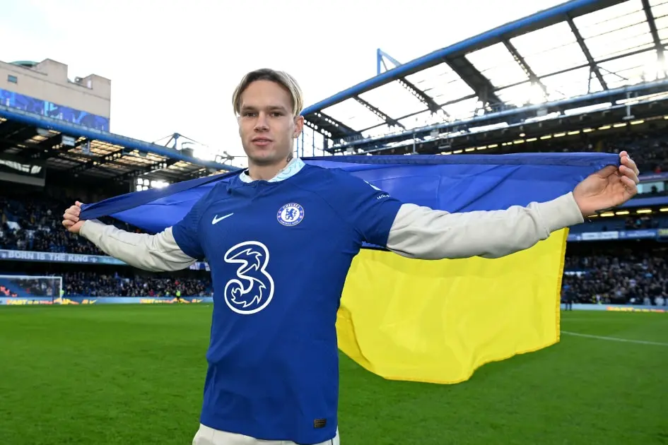 Mykhailo Mudryk wearing a Chelsea FC jersey and holding a Ukrainian flag at a stadium