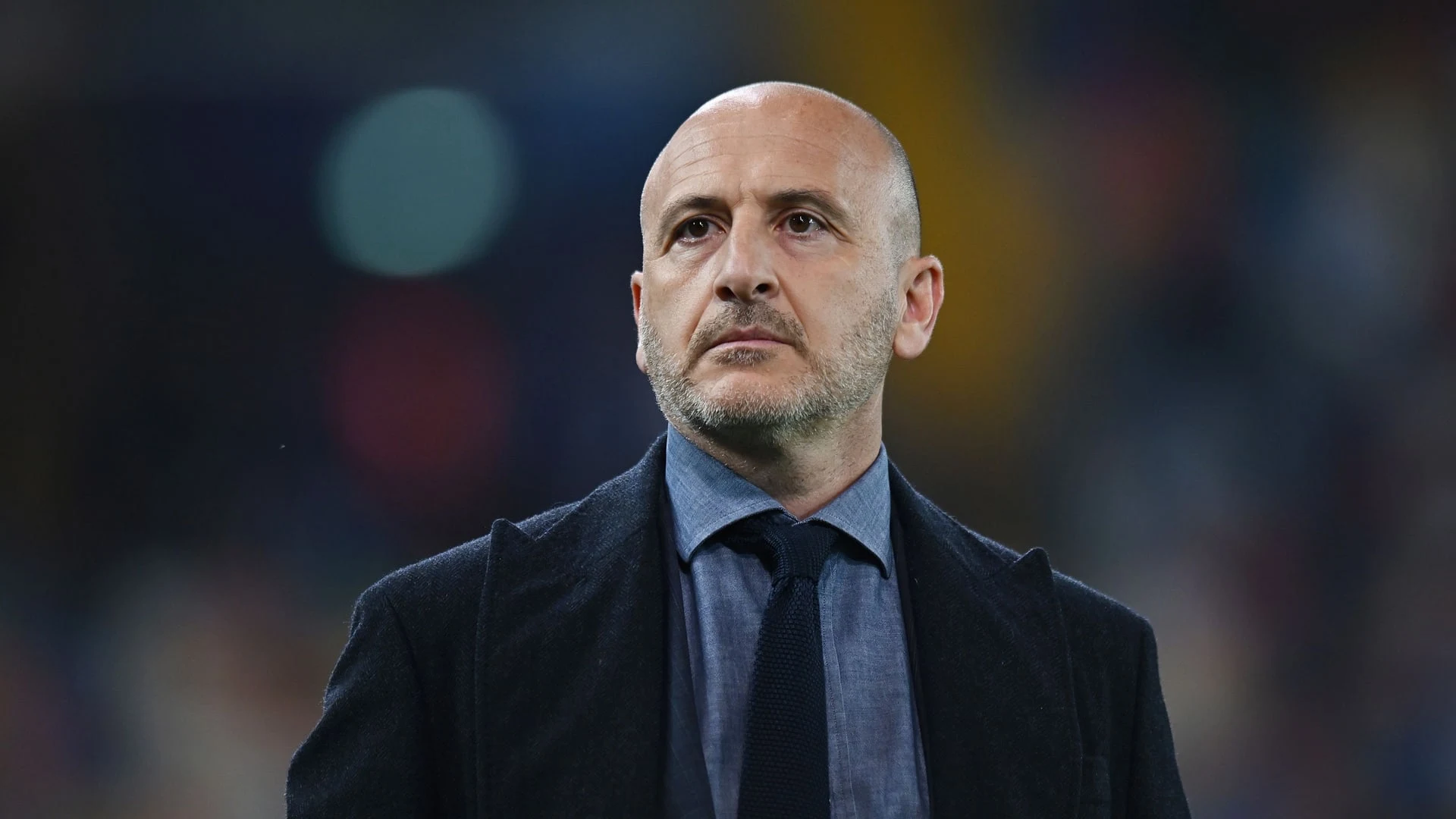 A bald man with a beard wearing a dark coat, blue shirt, and dark tie looking upwards.