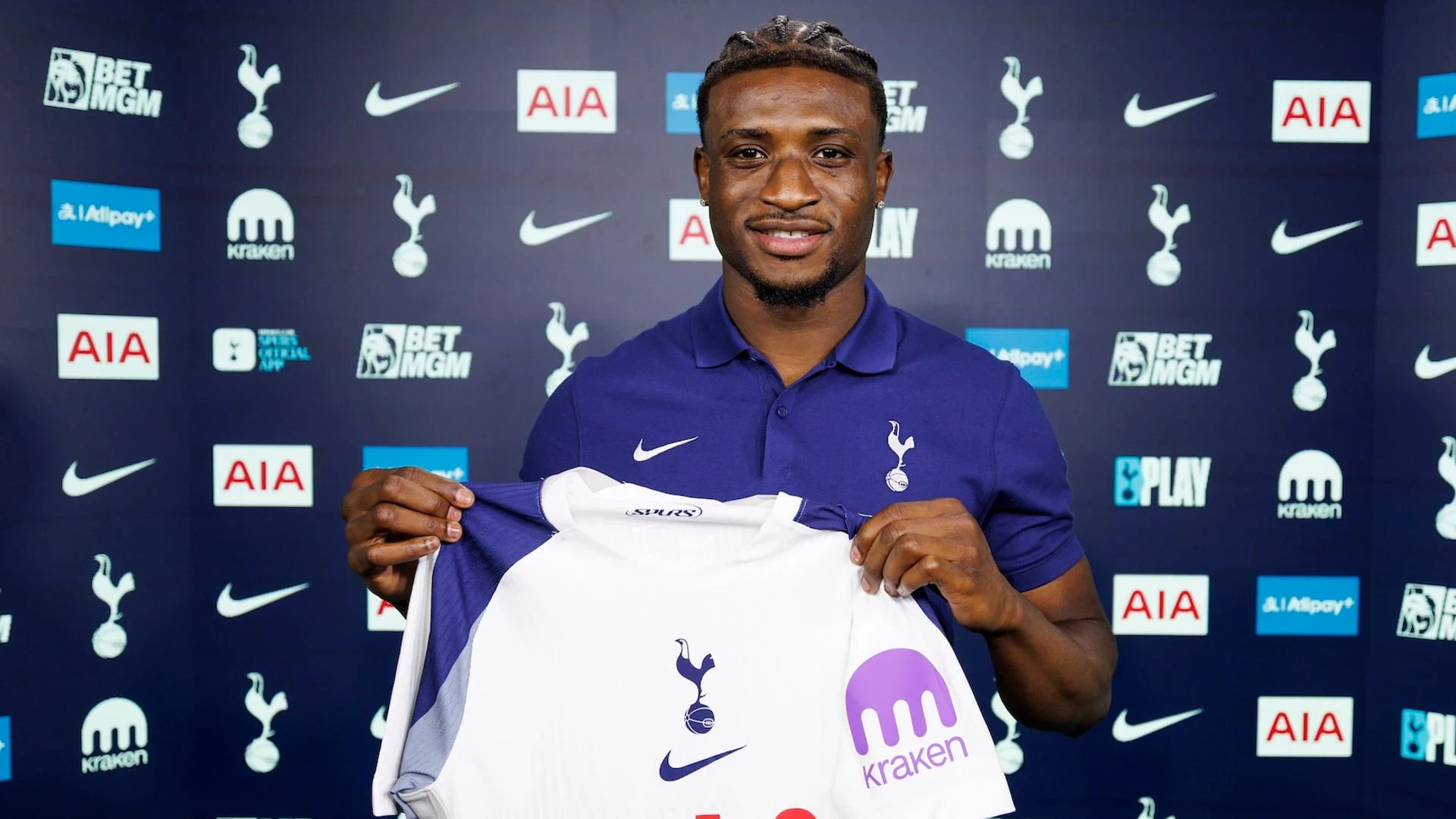 Mohammed Kudus smiling and holding up a Tottenham Hotspur jersey in front of a branded backdrop