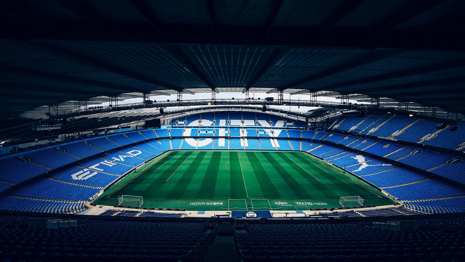 Wide view of the Etihad Stadium interior with blue seating and 'CITY' written across the stands.