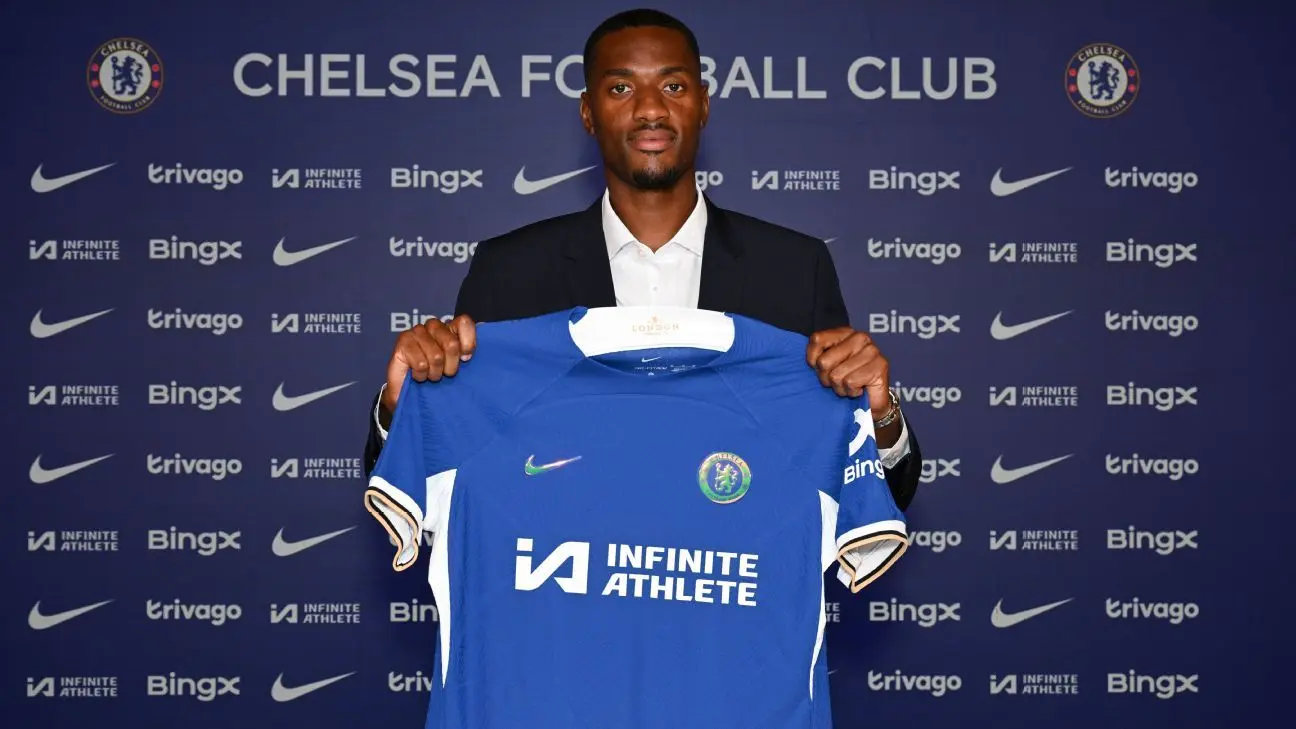 Tosin Adarabioyo posing in a suit while holding up a blue Chelsea FC home jersey