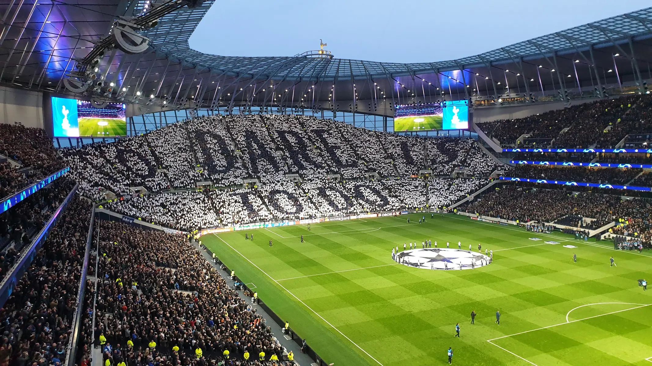 A wide shot of the Tottenham Hotspur Stadium with a large crowd in the south stand creating a tifo display.