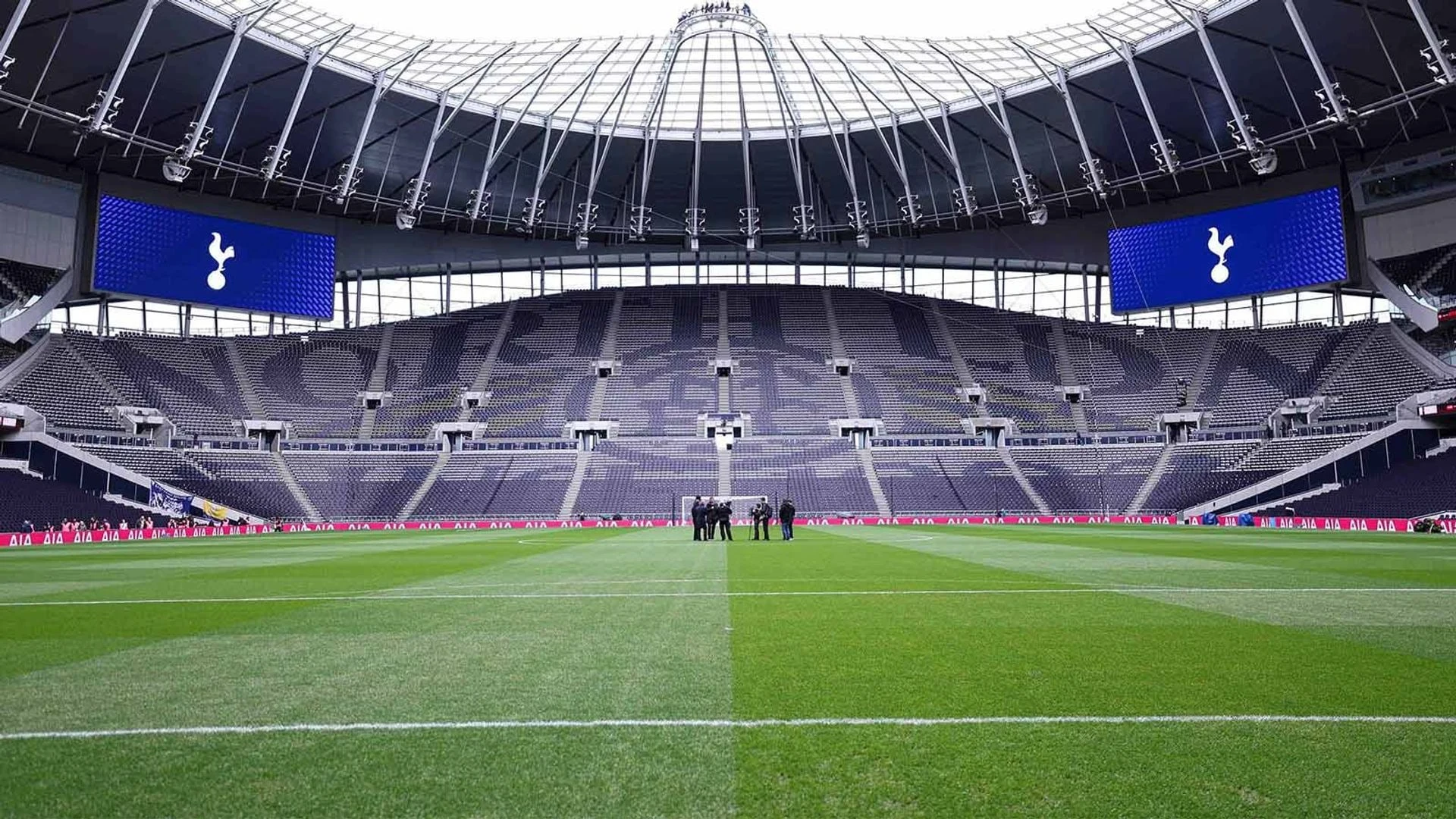 Wide view of the empty Tottenham Hotspur Stadium pitch and stands with club logos on the large screens.
